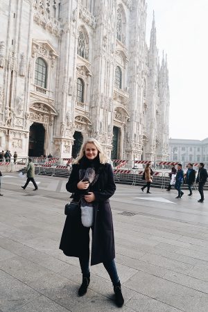 Lorna weightman wearing a long navy coat, jeans and boots in front of a large cathedral in Milan