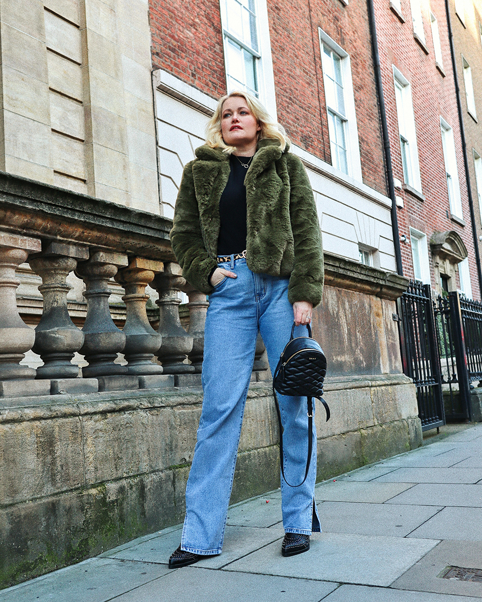 Lorna Weightman wearing green faux fur short jacket, black knitted jumper underneath, blue denim jeans with black ankle boots with studs. Holding a black bag