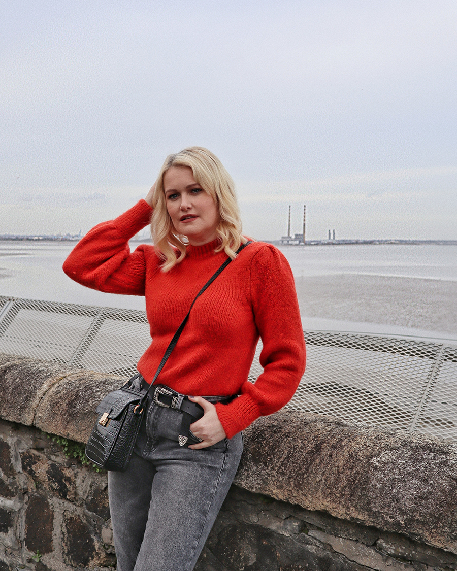 Lorna Weightman pictured in front of the sea wearing a red jumper and grey jeans holding a black bag